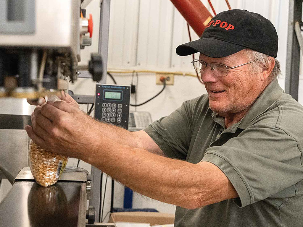 Person in baseball cap and glasses filling plastic bag with popcorn kernels from machine