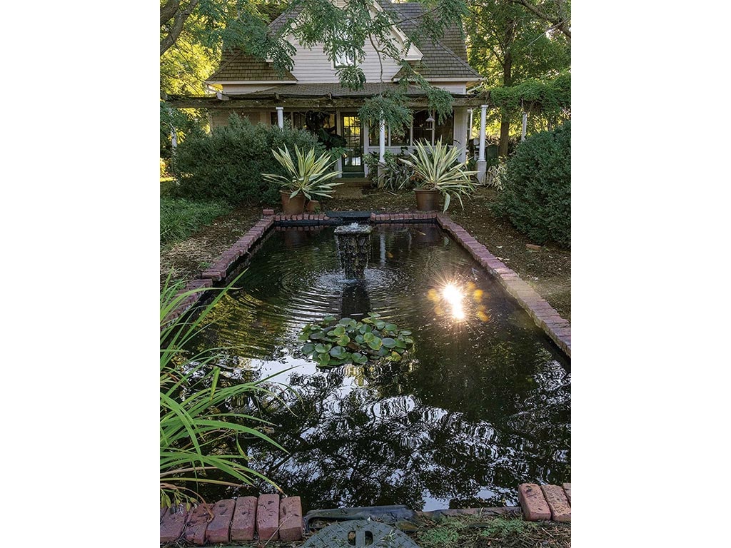 a pond with a fountain surrounded by landscaping in front of a wooded house