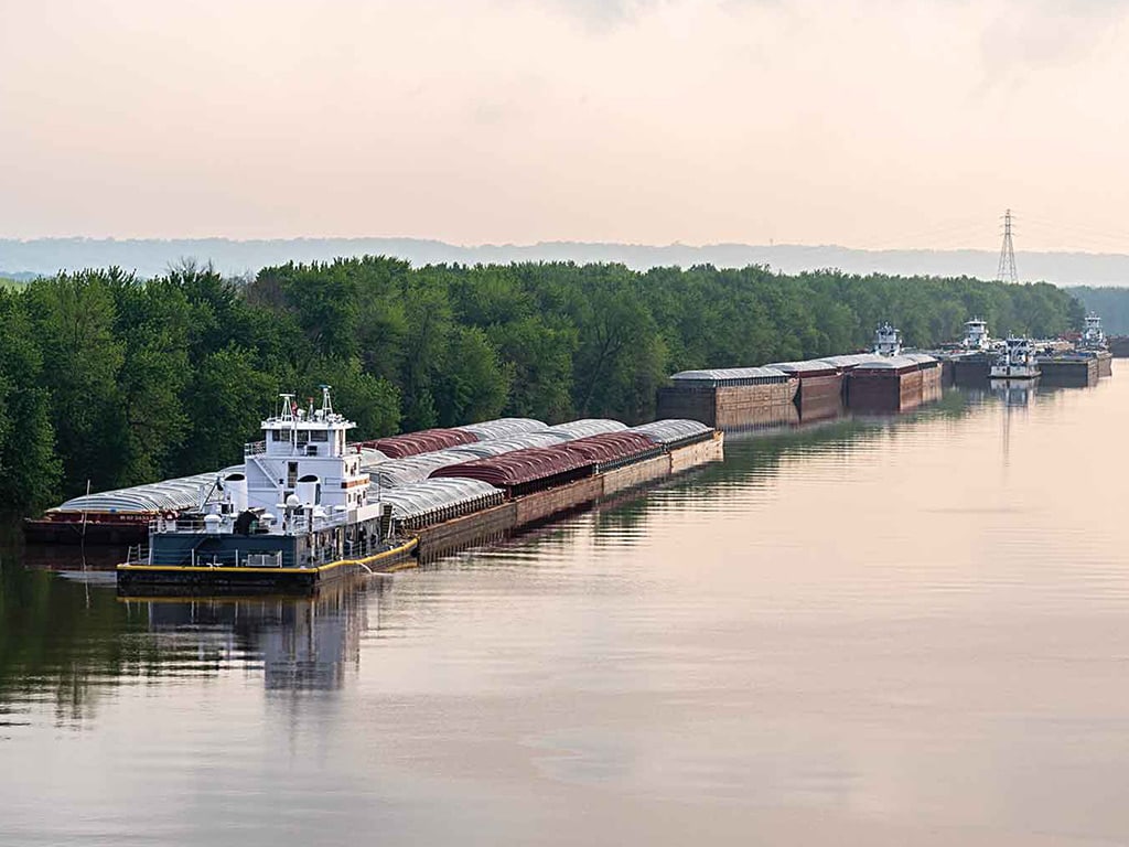 long view of a river shore with freight boats and barges backed by trees