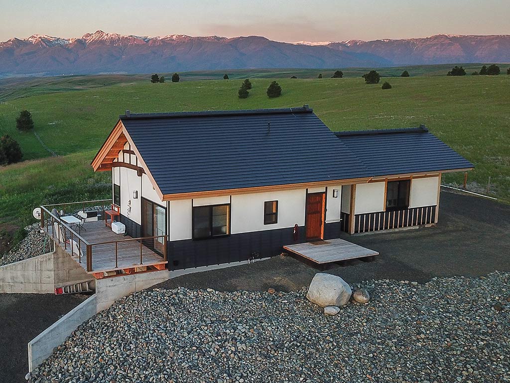 An aerial view of a Japanese style farmhouse with a large gravel yard in the foreground, a driveway leading under it, and green rolling hills leading out to a snowcapped mountain range in the distance