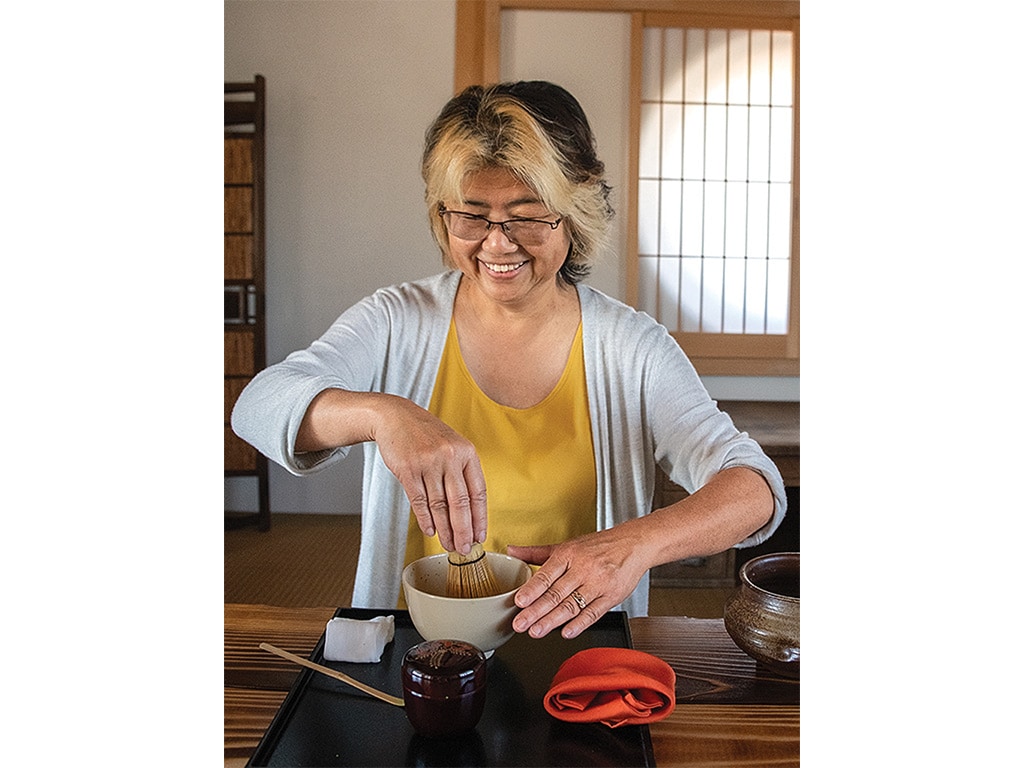 A smiling person traditionally whisking Matcha tea in a small stone bowl on a black serving tray on top of a wooden countertop