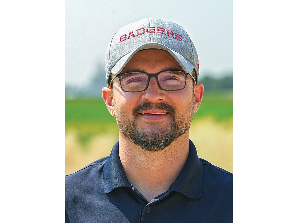 A man smiling with glasses and a hat on grass and crop field