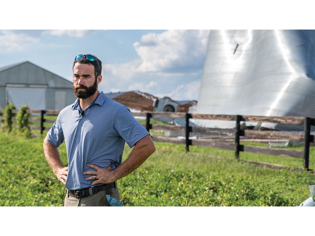 A farmer standing with their hands on their hips with a large dented shiny metal silo behind a fence in the background