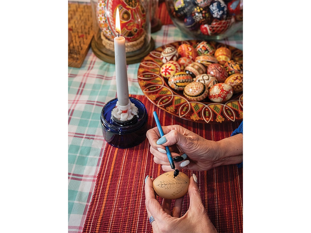 A closeup of a pair of hands applying wax to an egg, with a bowl of pysanky in the background.
