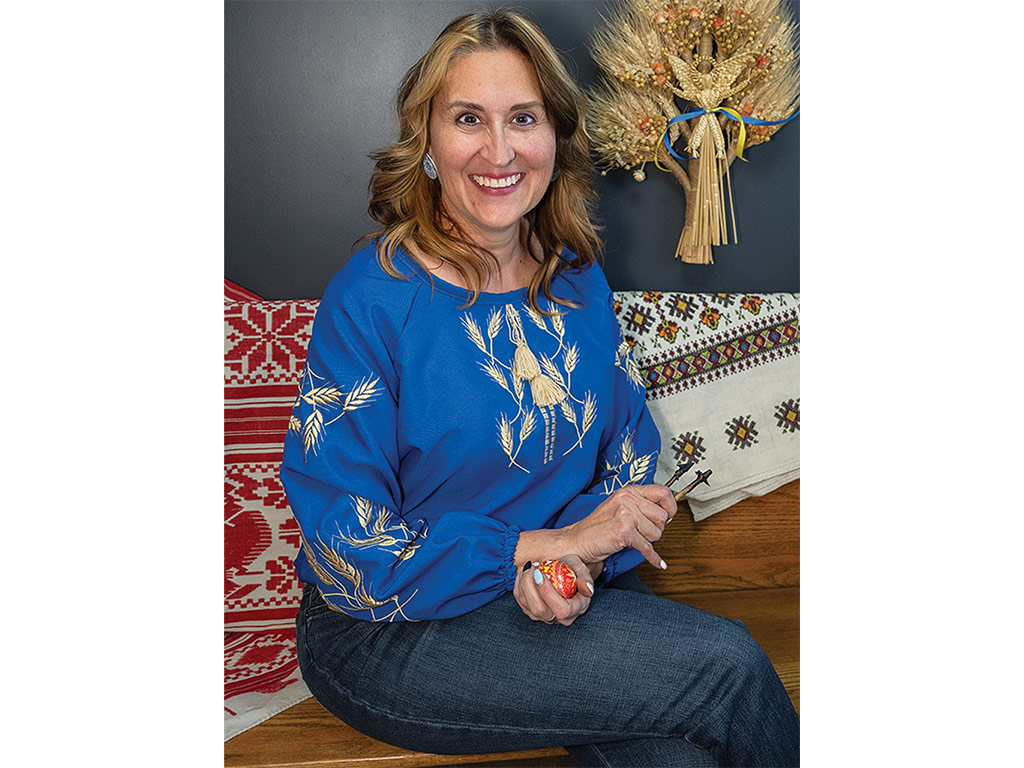 A seated woman wearing a blue wheat embroidered vyshyvanka shirt and holding a pysanky with a didukh woven wheat sculpture in the background.