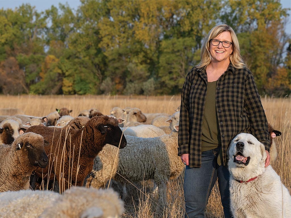 Perleberg smiling in a field with her multi-colored sheep flock on the left and a dog on the right.