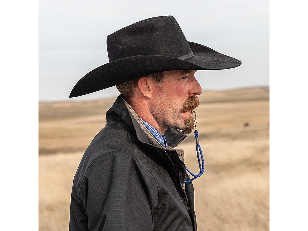 A closeup right-side view of a person in a field wearing a cowboy hat and gazing forward with a whistle in his mouth.