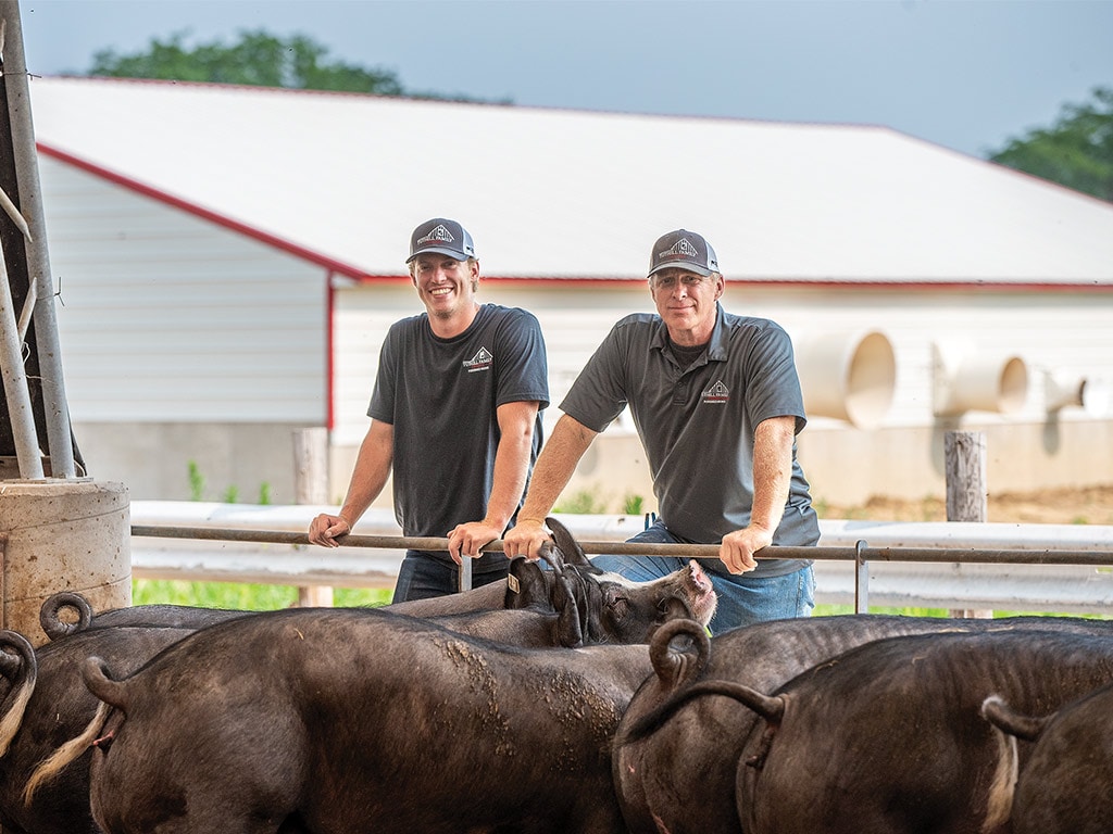 Two smiling pig farmers leaning on the rail of a pig pen