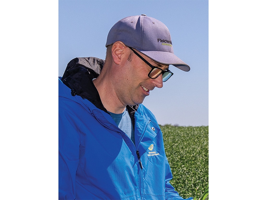Closeup of a person wearing a baseball cap standing in a field of crops, smiling and looking down