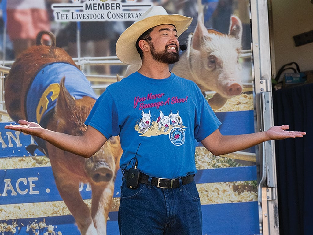 A man posing with his hands out in front of a pig racing poster, wearing a cowboy hat and a pig racing t-shirt
