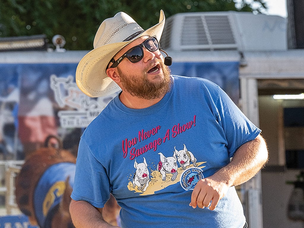 A man speaking into a mouthpiece, wearing a cowboy hat and a pig racing t-shirt