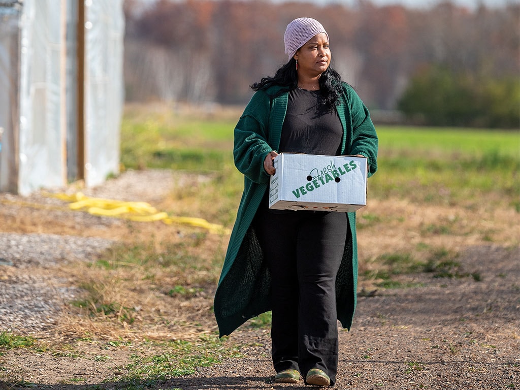 A farmer walking on a dirt path carrying a cardboard box of vegetables