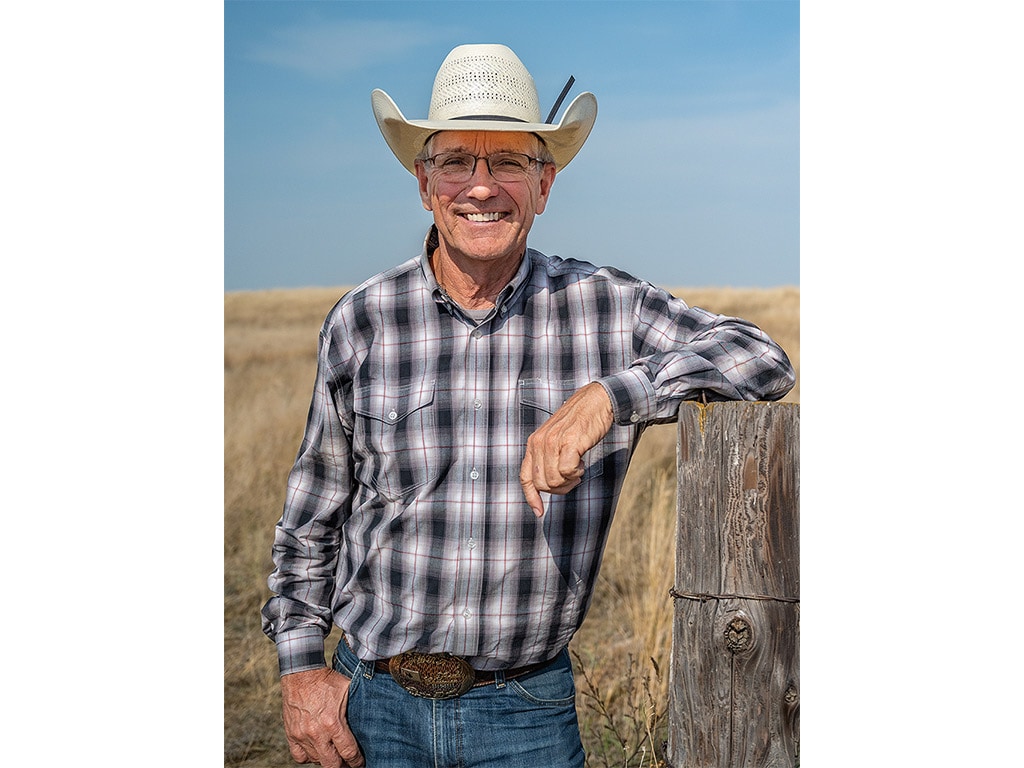 A smiling person with a cowboy hat leaning on a wooden post