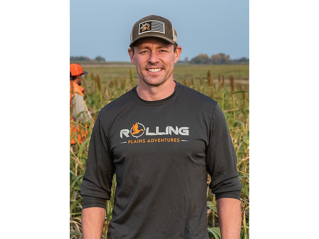 A smiling person with a baseball cap on standing in front of a field
