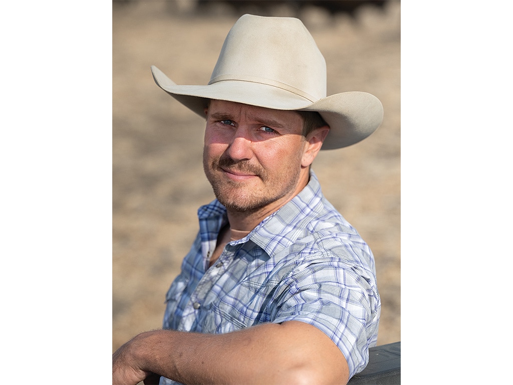 A smiling person with a cowboy hat leaning on a wooden post