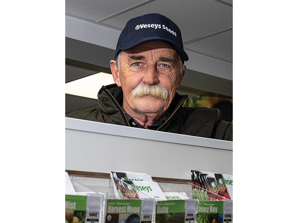A person with a white mustache and baseball cap looking over a retail store shelf with various seed packets displayed on it