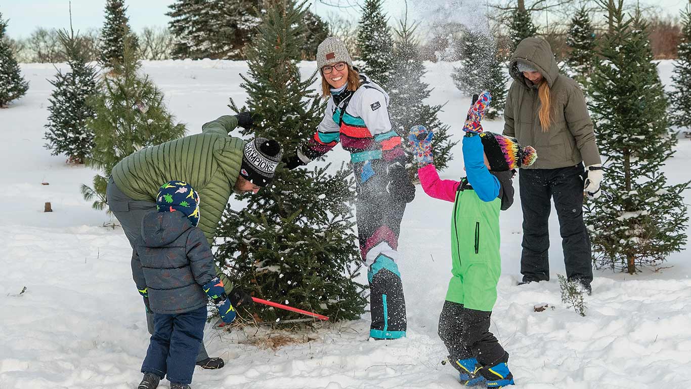 Family cutting a Christmas tree in a snowy field surrounded by evergreen trees.