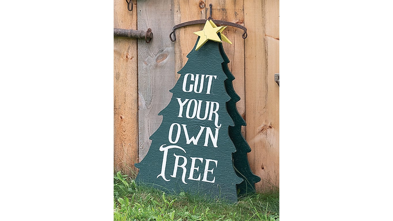 Christmas tree cutout sign reading "Cut your own tree" leaning against a wooden fence.