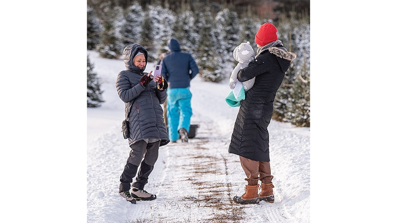 Mother holding child for a photo while another person pushes a sled at a Christmas tree farm.