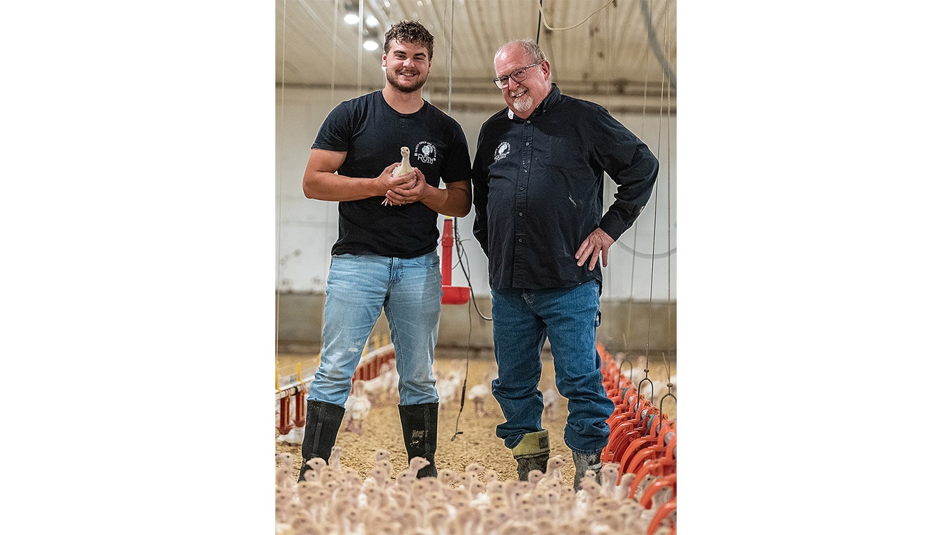 Two men stand in a poultry barn, one holding a young bird, surrounded by chicks on the ground.