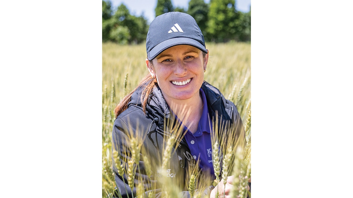 A person wearing a black jacket and a navy cap stands among golden wheat stalks in a sunny field.