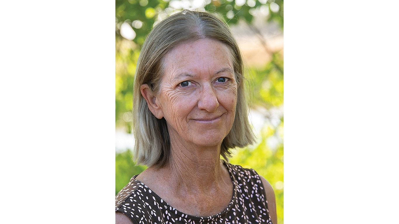 A woman with straight gray hair, stands outdoors surrounded by greenery.