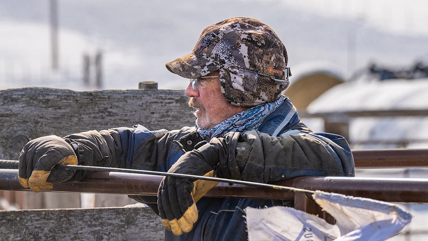 A person wearing a camouflage hat and gloves rests their arms on a wooden fence in a rural setting.