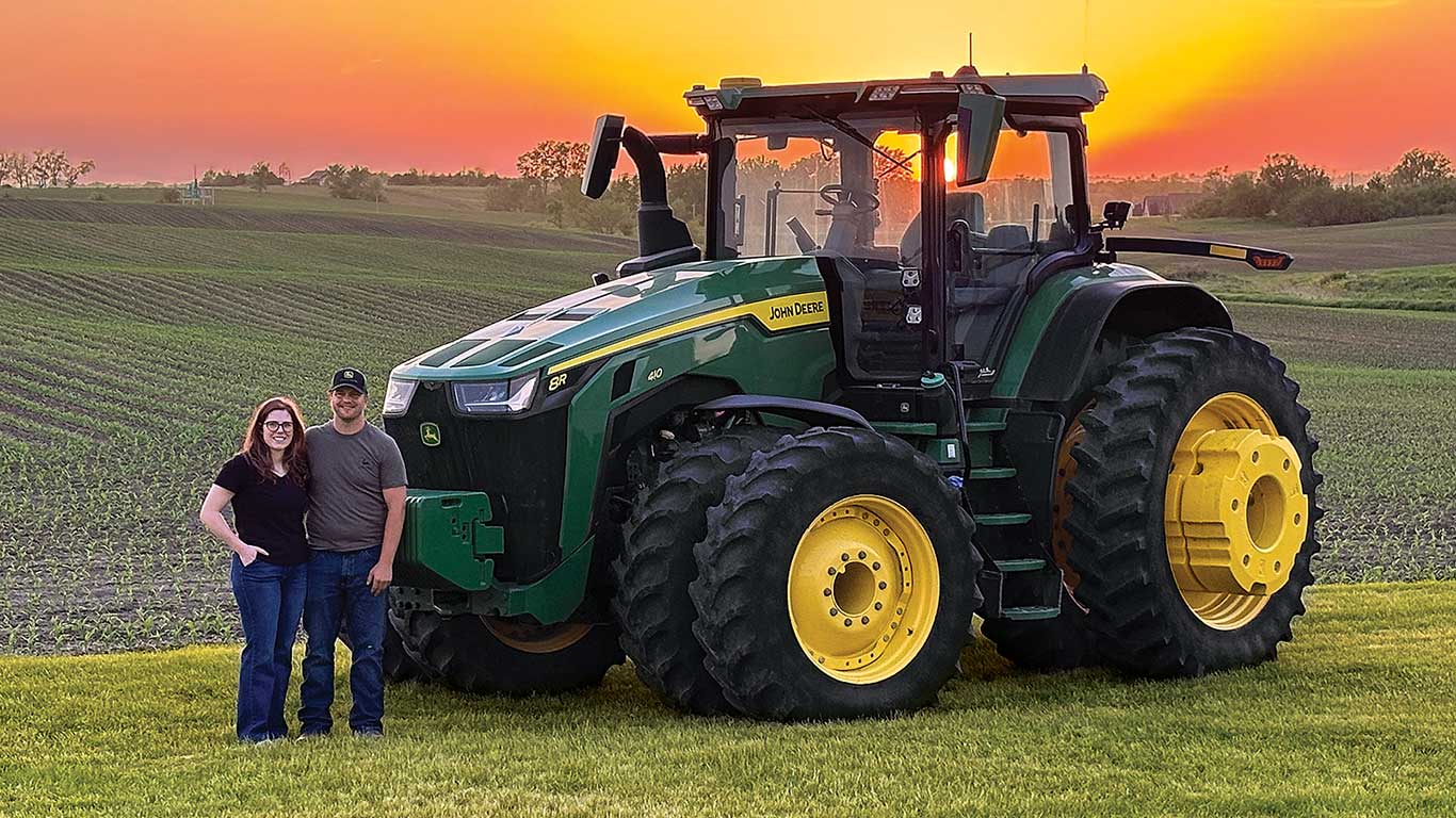 Green John Deere tractor on a grassy field at sunset with two people standing beside it.