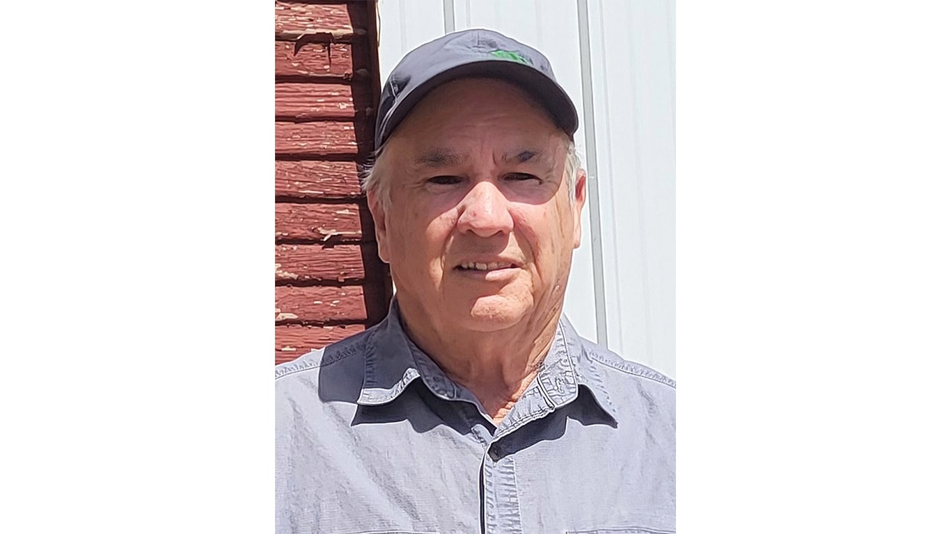 Person wearing a gray button-up shirt and cap standing near a red wooden wall and white siding.