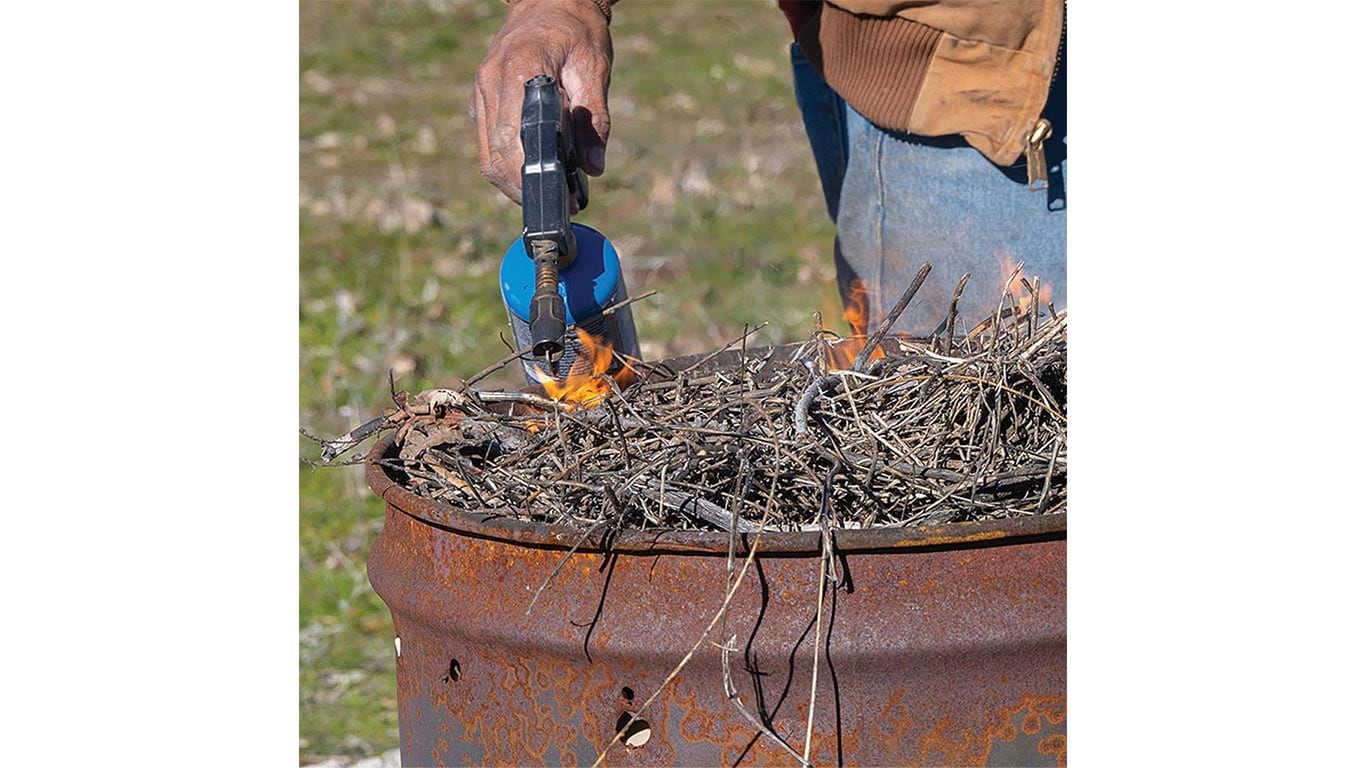 Hand holding blue torch lighting dry sticks in rusty barrel with small flames starting to appear.