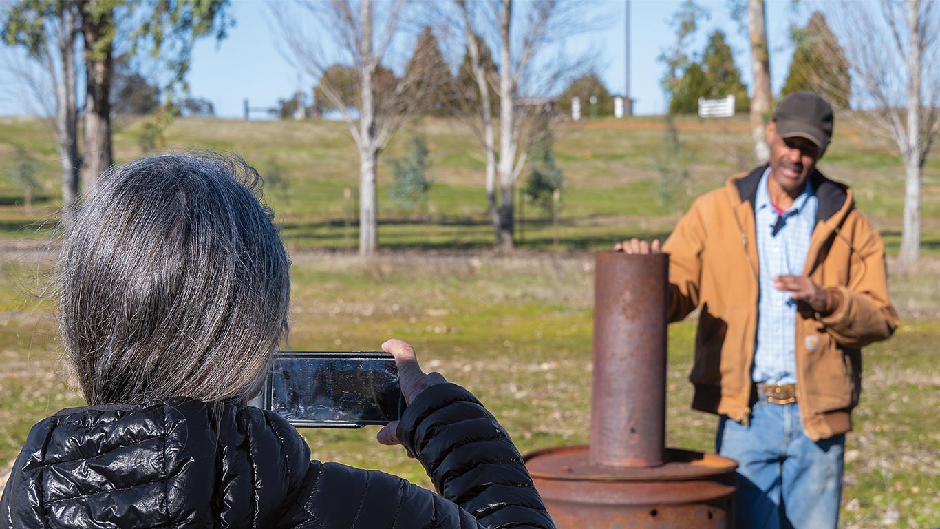 Person in black jacket photographing another by a rusty cylinder in a grassy outdoor area.
