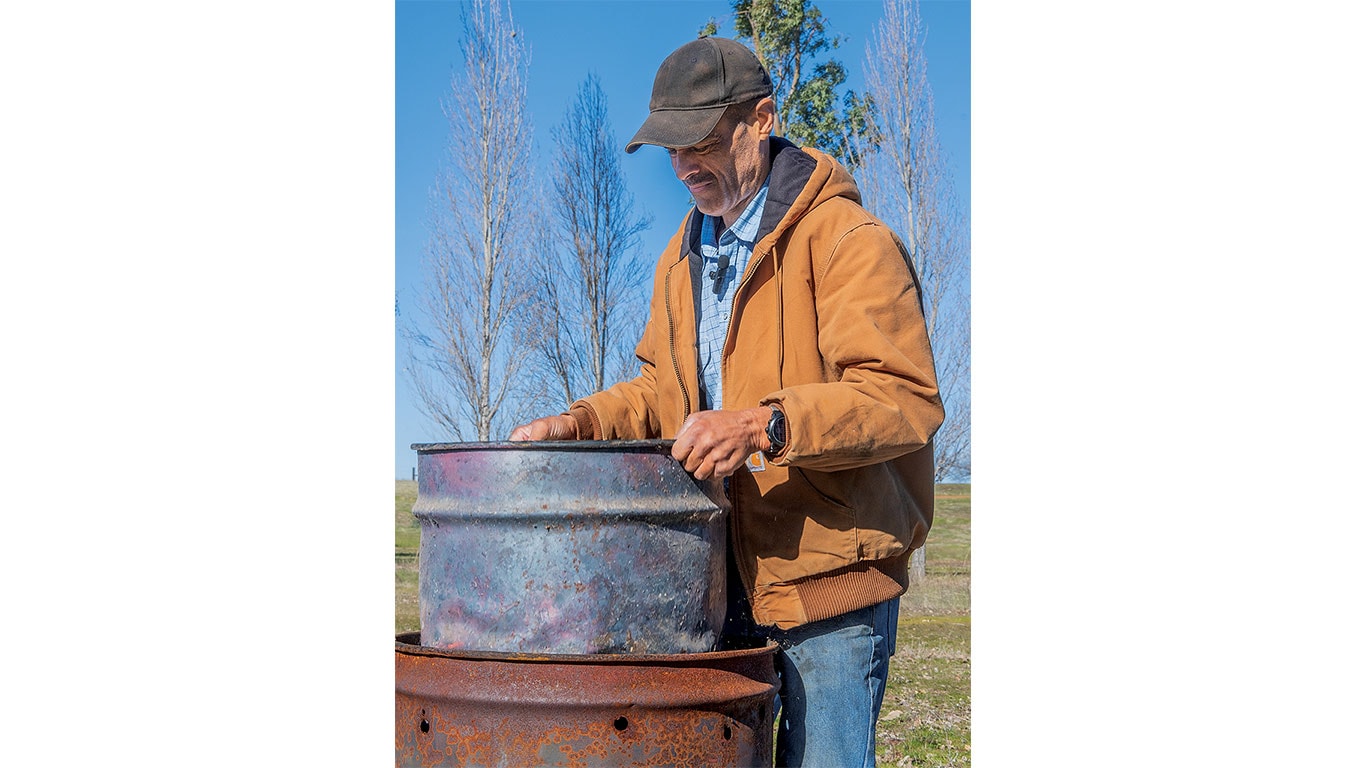 Person in brown jacket and cap handling metal barrel outdoors with leafless trees behind.