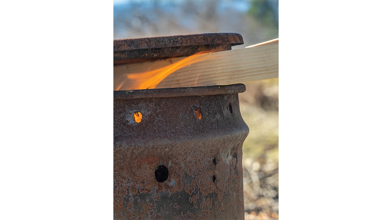 Rusty barrel with holes, burning fire inside, and a wooden plank sticking out from the top.