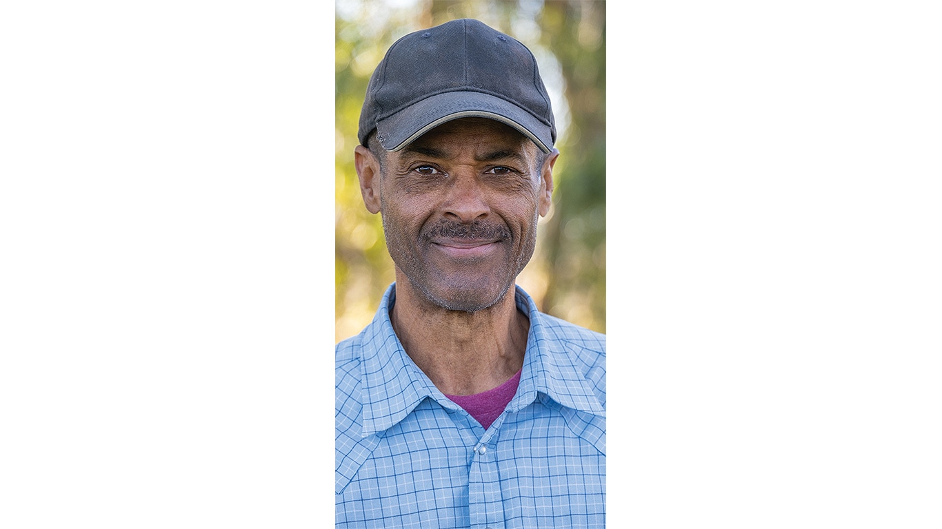 Smiling person in blue checkered shirt and dark cap, standing outdoors.