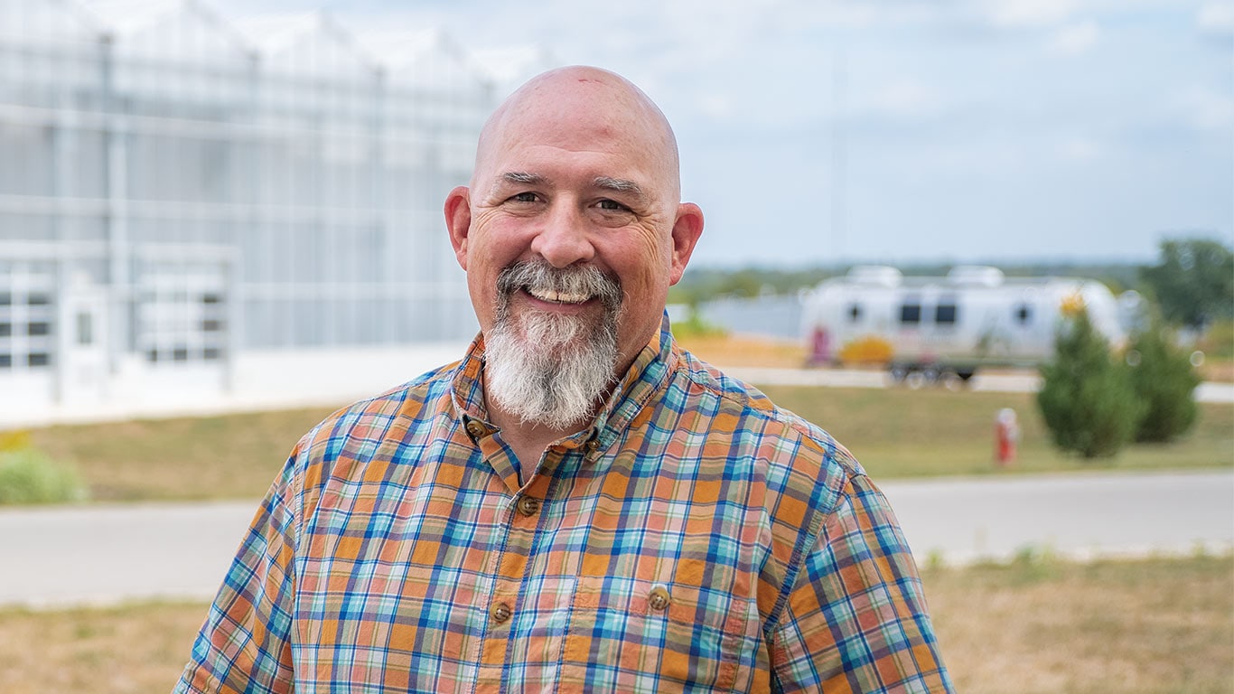 A man in a plaid shirt stands outdoors with a greenhouse in the background.