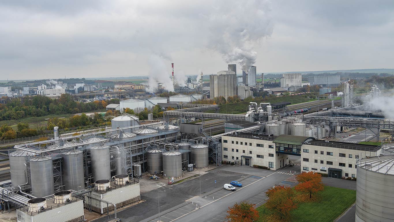 Aerial view of industrial site with storage tanks, buildings, and smoke emissions.
