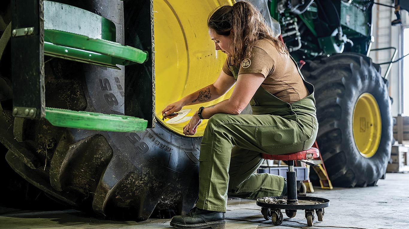 Person in green overalls measuring large tractor tire tread in workshop.