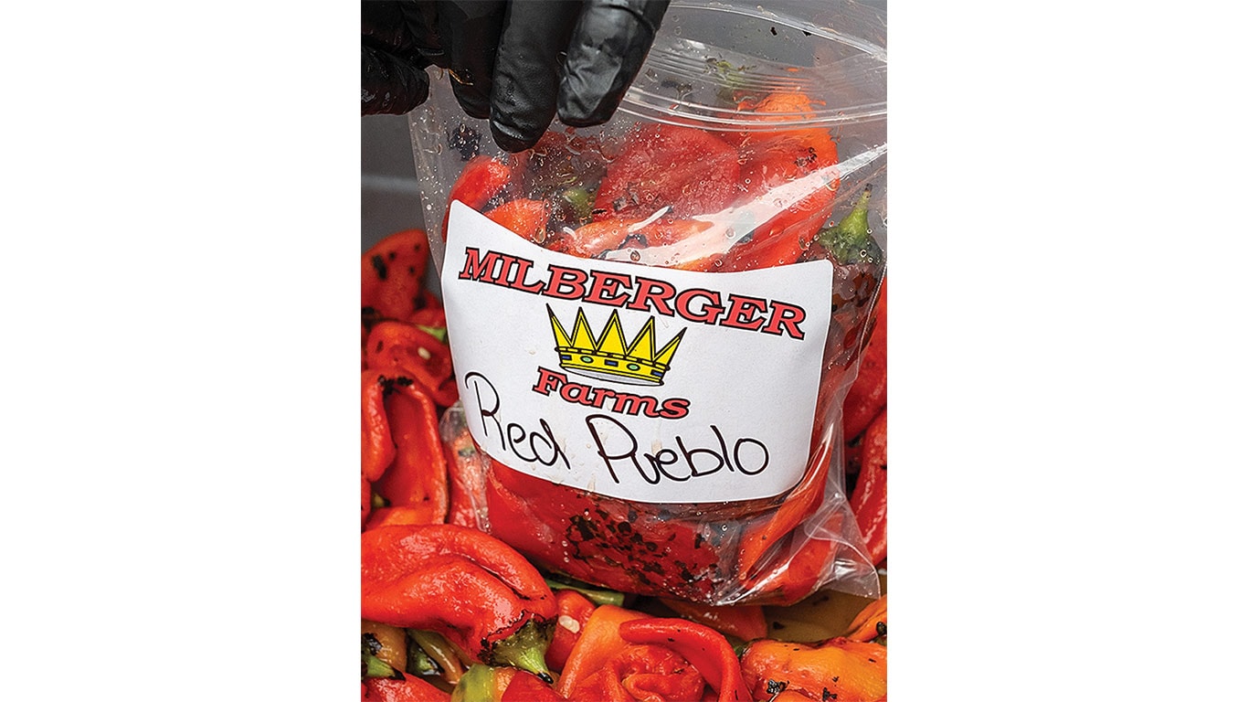Gloved hand holds bag of roasted red peppers labeled 'Milberger Farms Red Pueblo'.
