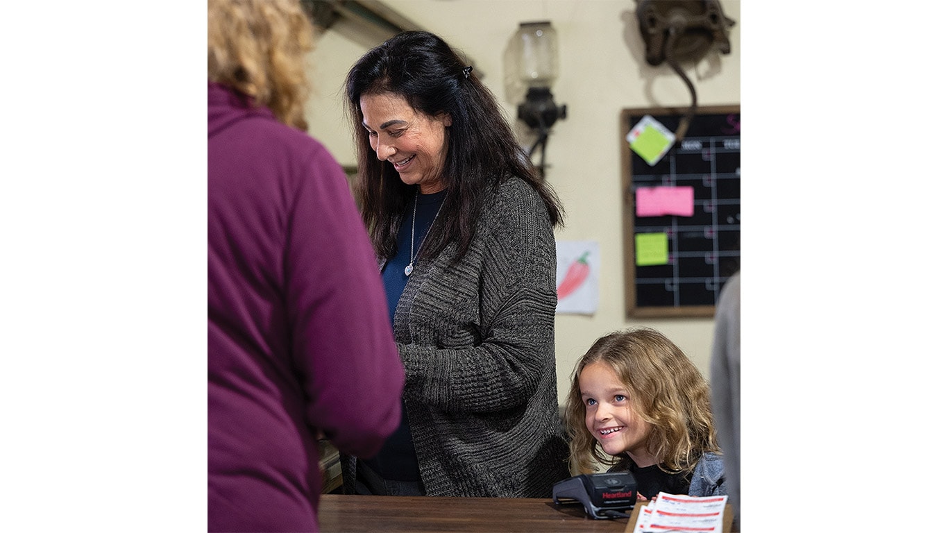 Adult in gray sweater and child with curly blonde hair stand at a counter indoors.