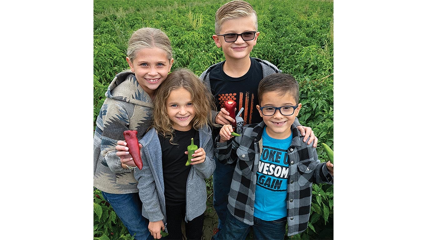 Five children stand in a green field, each holding a different vegetable.