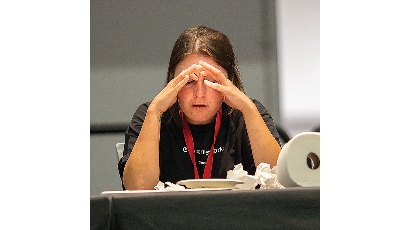 A person with long hair sits at a table, resting their forehead on clasped hands, with a plate and tissues nearby.