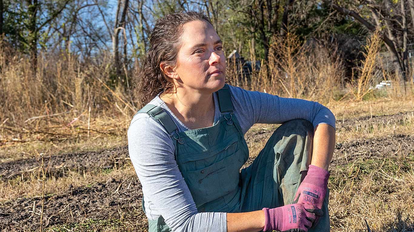 Person in overalls and gloves sits in garden beside tray of seedlings and tools.