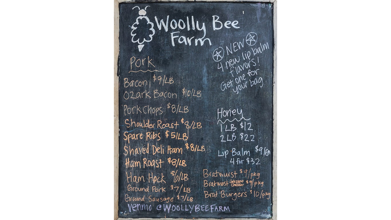 Chalkboard menu at Woolly Bee Farm listing pork, honey, lip balm, and bratwurst with prices.