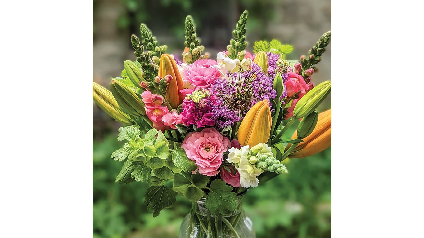 Bouquet with pink roses, orange lilies, and green leaves in a glass vase, greenery background.