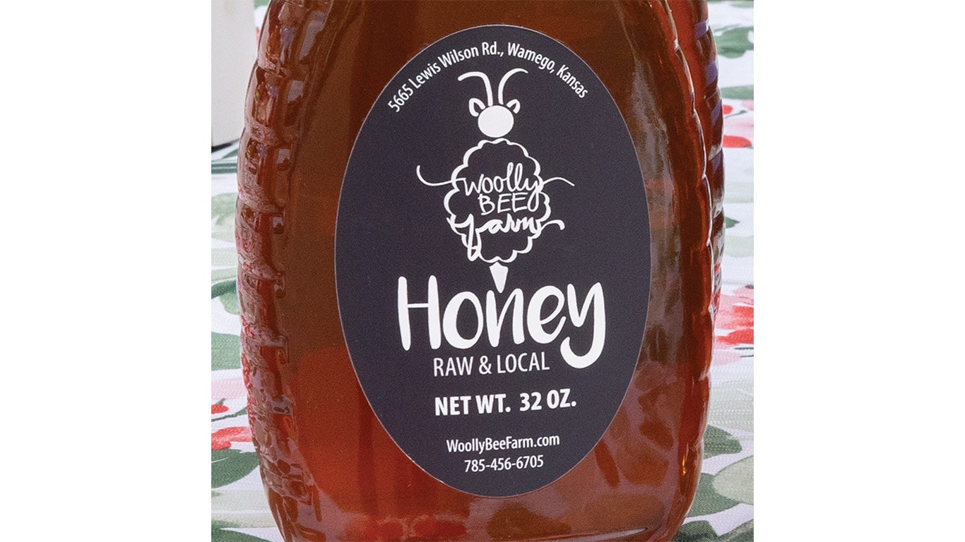 Jar of Woolly Bee Farm honey with black label showing address, website, and phone number.