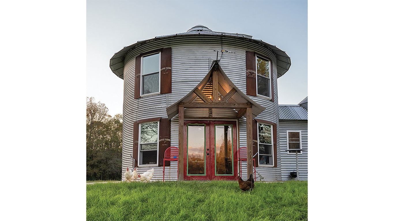 Round metal house with red doors, wooden porch, and chickens on grass with trees in background