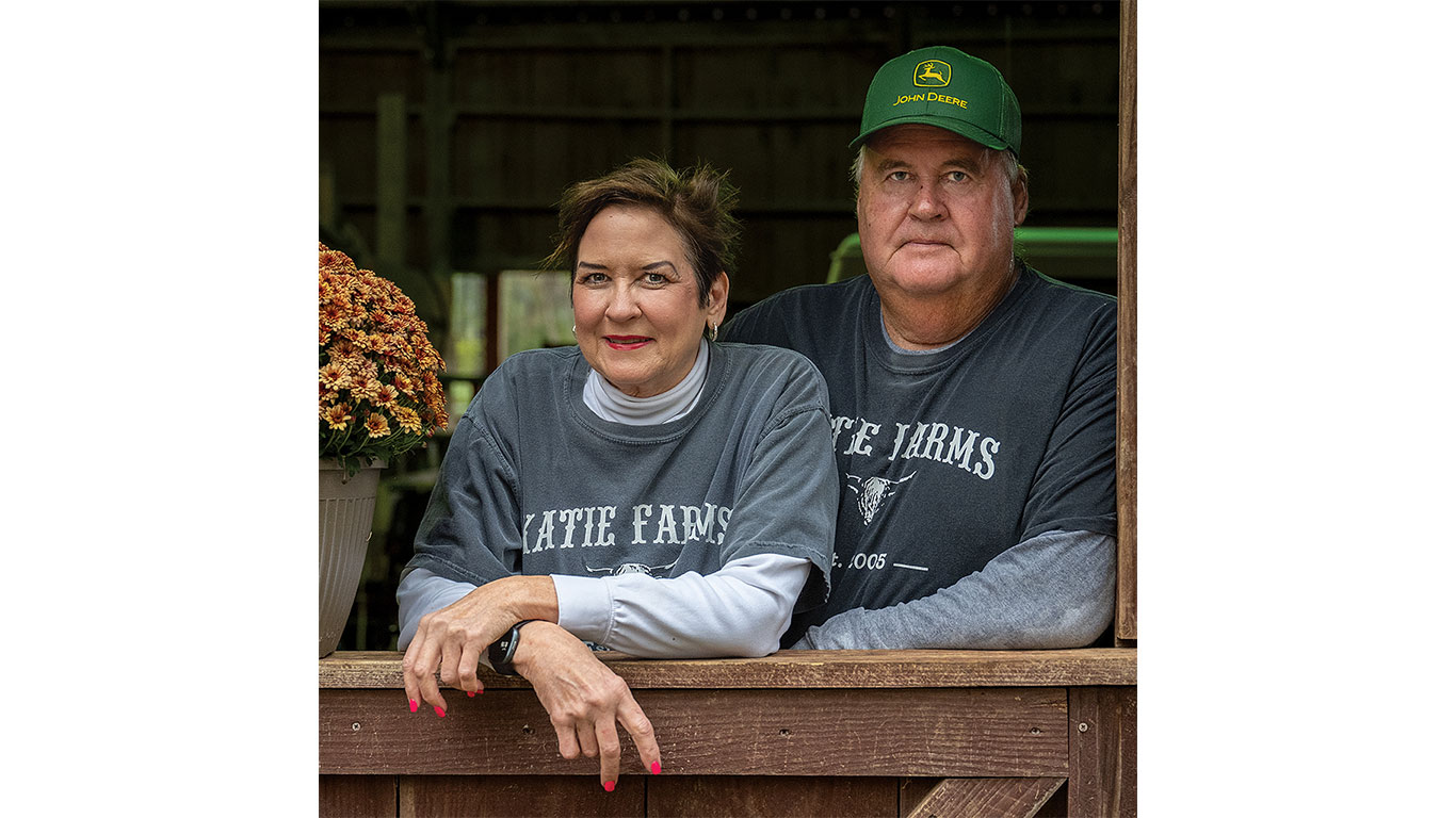 Two people in Katie Farms shirts lean on fence with a flower pot and green cap visible