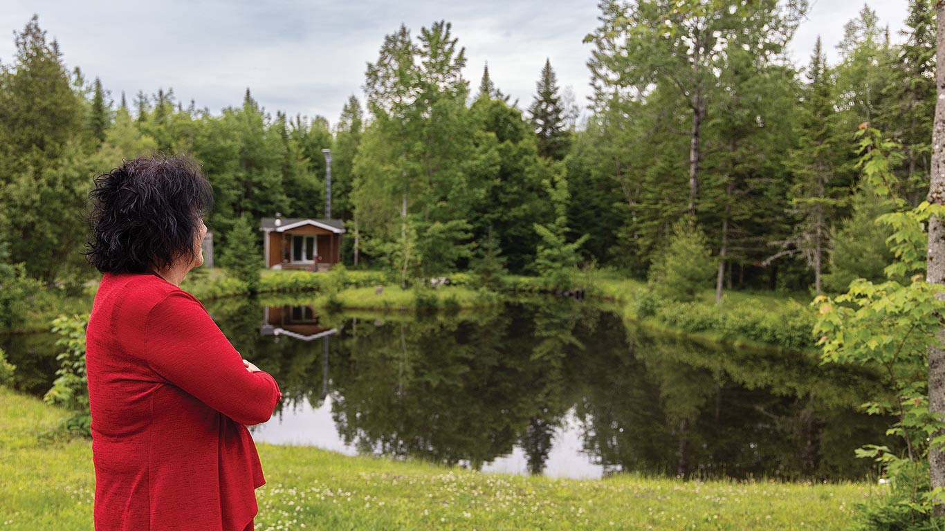 Person in red jacket looks at pond with dense green trees and small cabin in background.