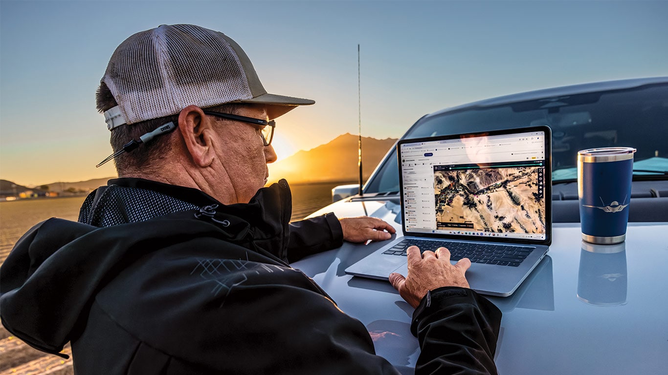 Person uses laptop with map on vehicle hood, cup nearby, mountains and sunset in background.