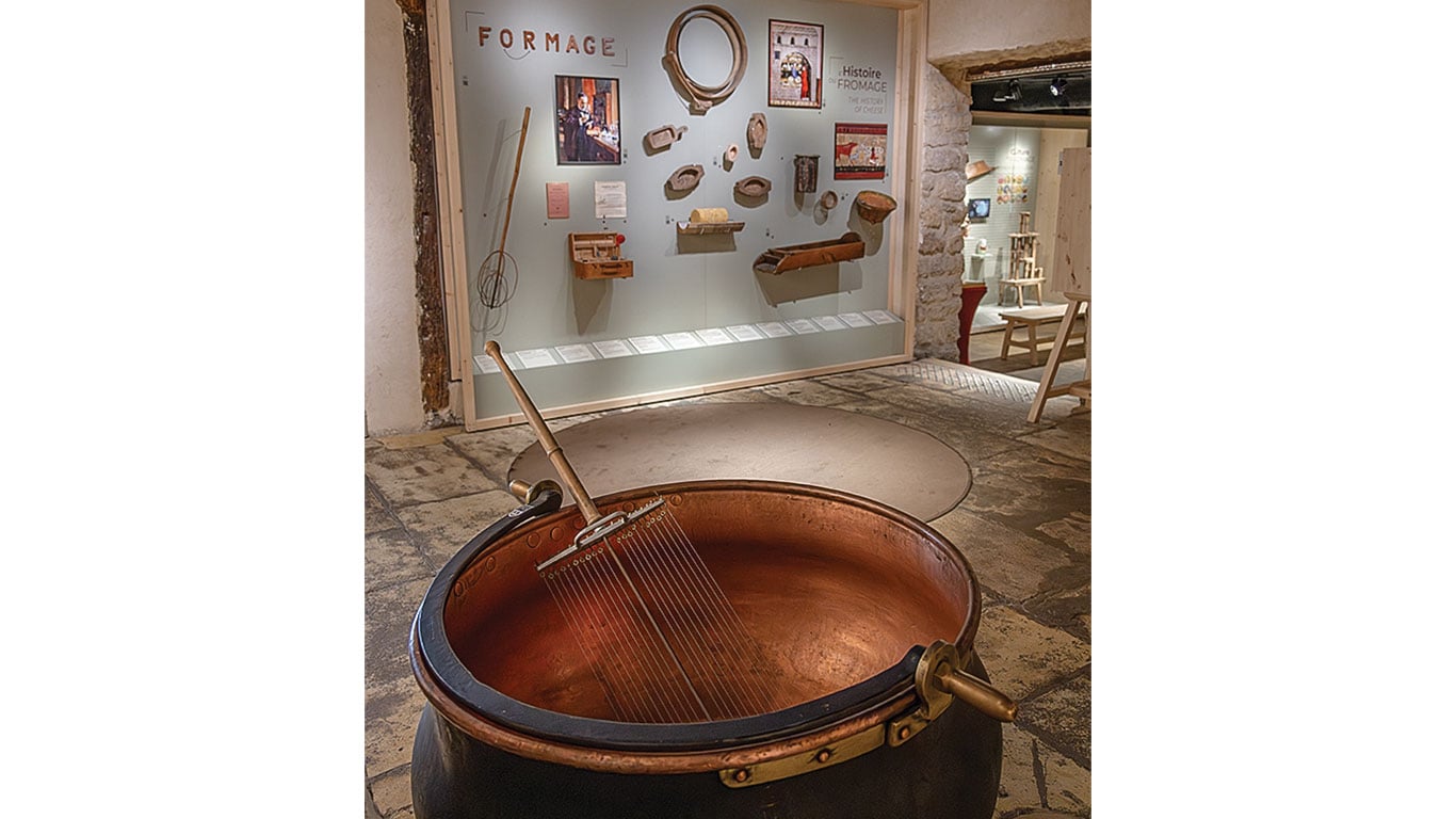 Copper cheese vat with cutting tool in front of a museum display on cheese-making history.